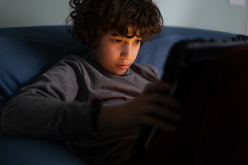 Lille boy watching video contents or playing video games holding a tablet computer on the legs during the night, his face is lighted by the colored screen light. Child addicted by internet technology.