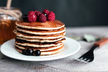 Juicy pancakes with berries and honey on a white plate, spoon, jar, wooden table