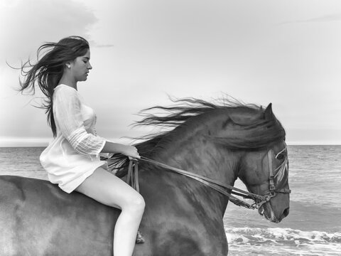 Joven amazona con vestido blanco domando a su caballo marron sin montura en la playa