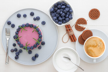 The two-color blueberry mini cheese cake. Round no bake cheesecake, cut portion on small plate with few cookies in the focus. The top of cake decorated by rosemary herbs. White table.