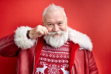 grey-haired santa man in coat touching mustache isolated on red background, portrait of senior man...