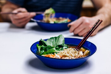 Ramen noodles with egg and bok choy in a cobalt blue bowl, man eating in the background