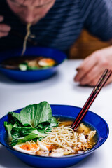 Ramen noodles with egg and bok choy in a cobalt blue bowl, man eating in the background