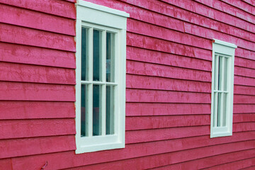 Fototapeta na wymiar Two vintage double hung windows with six panes of clear glass in each, on the exterior of a bright red wall of worn wooden horizontal clapboard. The thick trim on the windows is stark white. 