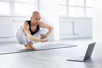 Bald man performing complex yoga pose through online instructions, young caucasian guy recording video on laptop, sit in bright modern studio, training alone. during coronavirus quarantine