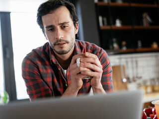 Handsome man preparing breakfast at home. Young man drinking coffee and using the laptop.