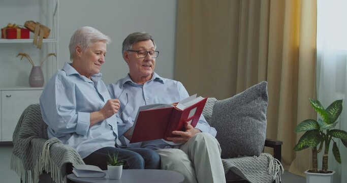 Old couple Caucasian elderly family gray-haired man and woman grandparents sitting on sofa in cozy living room looking in photo album remembering pleasant moments of life enjoying memories, nostalgia