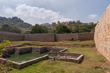 Chitradurga, Karnataka, India - November 10, 2013: Fort or Elusuttina Kote. Small green water tank surrounded by brown stone wall under blue cloudscape with boulder hills and trees in between.