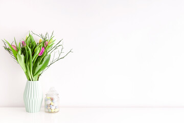 Bouquet of multicolored unopened tulips in light green ceramic vase and Easter candy jar filled with eggs on white wall background. Home interior decoration concept. Place for text.
