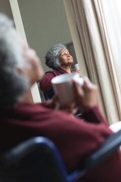 Thoughtful african american senior woman holding coffee cup looking out of window while sitting on w