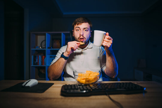 Concentrated Man Sitting At Night At The Computer Desk And Eating Chips And Drinking Tea From A Cup, Looking At The Screen With A Serious Face. The Programmer Works At Home At Night And Eats Chips.