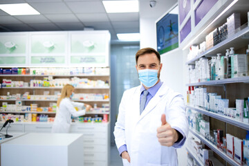 Portrait of pharmacist wearing face mask and white coat showing thumbs up in pharmacy store during...