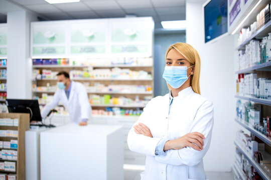 Portrait of female pharmacist wearing face mask and white coat standing in pharmacy store during corona virus pandemic.