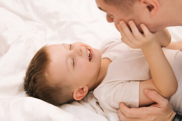 Fototapeta na wymiar Young dad playing and laughing with his baby smiling son at home in bed in the bedroom, happy family