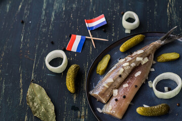 Traditional Dutch food herring fish with pickled cornichons and onion on dark blue ceramic plate and wooden background with flags. New season of herring called "Hollandse nieuwe haring" .copy space. © Tamilla