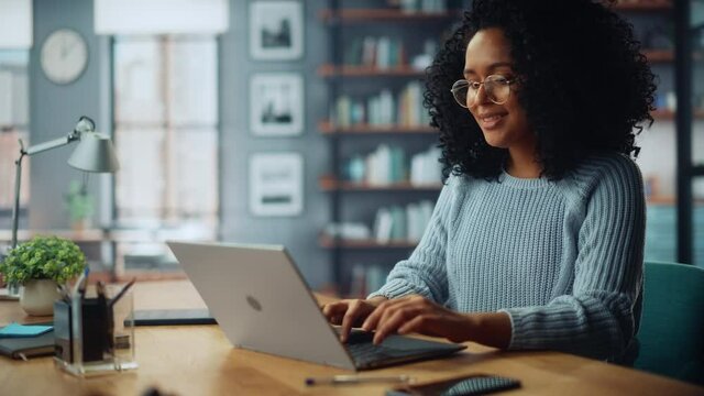 Beautiful Authentic Latina Female Sitting at a Desk in a Cozy Living Room and Using Laptop Computer at Home. She's Browsing the Internet and Checking Videos on Social Networks. Having Fun at Home.