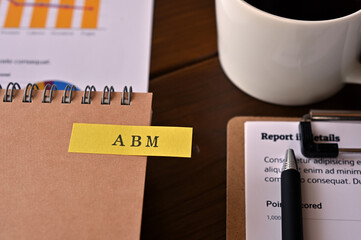 There's a cup of coffee, a laptop and a notebook with a sticky note on it that says ABM written on...