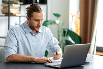 Concentrated caucasian guy freelancer or student uses laptop, distant learners or work. Focused attractive modern man in stylish casual wear sits at the desk in a creative or home office, browsing