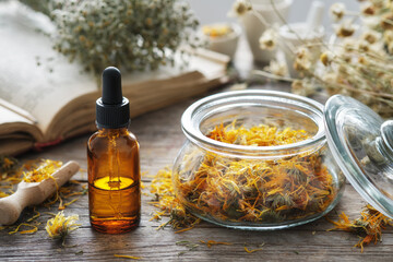 Dropper bottle of calendula infusion or oil, jar of dried marigold flowers, old recipes book and chamomile bunch on background. Alternative medicine. - obrazy, fototapety, plakaty