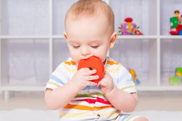 Cute caucasian baby boy playing with wooden toys. Sitting on white carpet in striped bodysuit. Different children's toys in the background. Looking down. Red wooden toy in mouth.