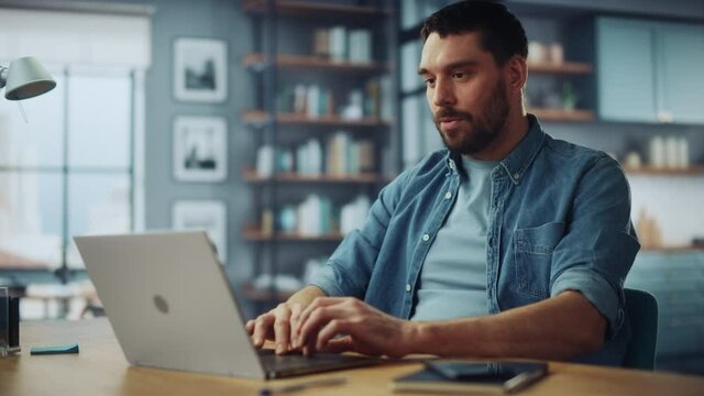 Handsome Caucasian Man Talking on Video Call on Laptop Computer while Sitting on a Sofa in Stylish Living Room. Freelancer Working From Home. Browsing Internet, Drinking Coffee from a Mug, Having Fun.