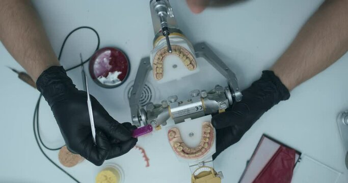 A dental technician is wearing black disposable gloves, holding an articulator with an upper and a lower jaw in it, and applying some white mass onto one of the teeth and then wiping it with a brush.