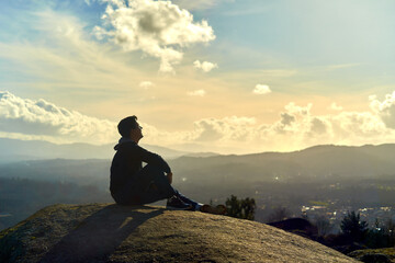 young boy sitting on top of a mountain looking to the horizon. enterprising man sitting on a rock with background sunset landscape. dark teenager with Latino features on a mountain