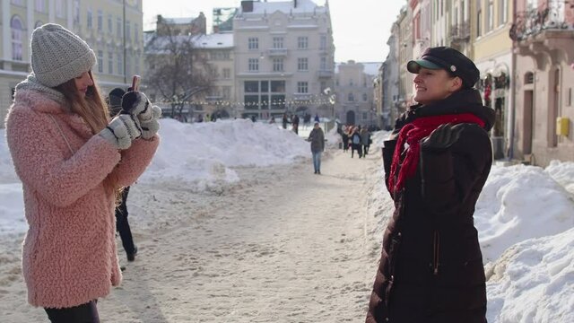 Happy lesbian LGBT couple during winter holidays traveling vacation making online vlog. Two young smiling women tourists bloggers taking photos portrait on mobile phone while standing on city street