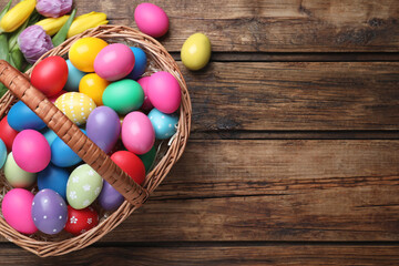 Fototapeta na wymiar Colorful Easter eggs in wicker basket and tulips on wooden table, flat lay. Space for text