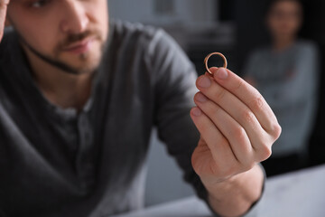 Sad man holding wedding ring at table indoors, closeup. Couple on verge of divorce