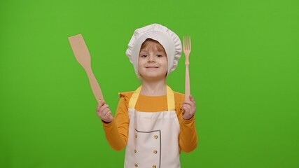 Fototapeta premium Child girl kid dressed as professional cook chef showing wooden fork and spatula, smiling, nods head in agreement on chroma key background. Nutrition, cooking school, education, food. Fun and humor