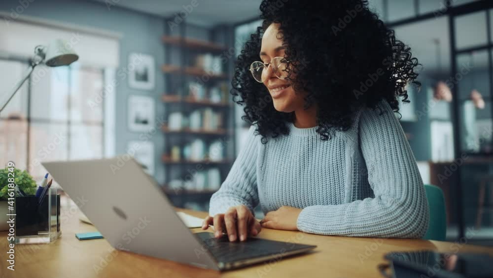 Poster beautiful authentic latina female with afro hair sitting at a desk in a cozy living room and using l