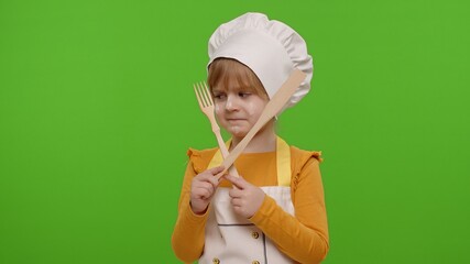 Dissatisfied child girl kid dressed as professional cook chef with wooden fork and spatula, showing...