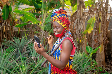 A beautiful happy African female farmer with nose mask, holding a smartphone with a hoe hanging from her shoulder in her pineapple and banana farm  