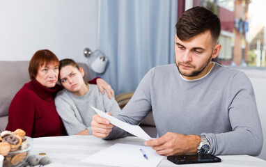 Frustrated young man looking at papers on background with discontented wife and her mother