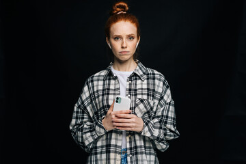 Doubtful sad red-haired young woman wearing wireless earphones typing online message on isolated black background, looking at camera. Pretty redhead lady model emotionally showing facial expressions.