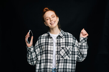 Positive young woman wearing wireless earphones holding phone and dancing listening online music on isolated black background. Pretty redhead lady model emotionally showing facial expressions