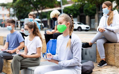 Students in protective masks record lecture while sitting on a stone street parapet