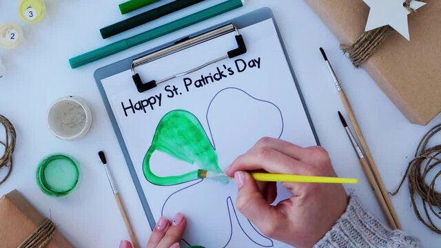 Female hands painting greeting card St Patricks day. Gift idea, decor craft. DIY. Do it yourself. Clovers, brush and paint. Saint Patricks Day hat.