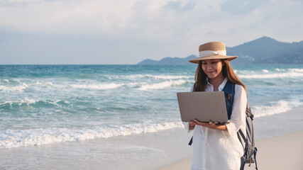 A young female traveler using and working on laptop computer while walking on the beach