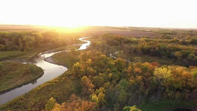Drone shot over creek surrounded by fall colored trees during sunset