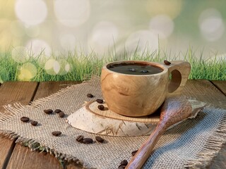 An oak table with visible grains is standing on a wood cup with black coffee. On a jute napkin, coffee beans are scattered on a birch pad 