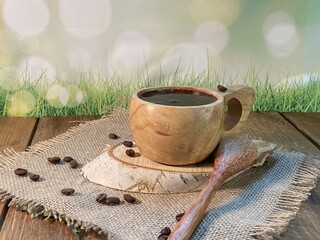 An oak table with visible grains is standing on a wood cup with black coffee. On a jute napkin, coffee beans are scattered on a birch pad 