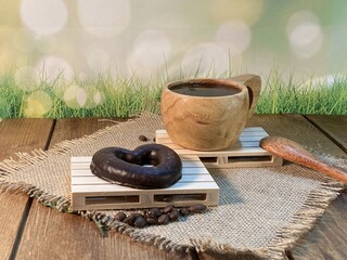 An oak table with visible grains is standing on a wood cup with black coffee. On a jute napkin, coffee beans are scattered and a gingerbread bitten heart lies on the pallet  © Józef