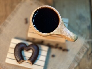 An oak table with visible grains is standing on a wood cup with black coffee. On a jute napkin, coffee beans are scattered and a gingerbread bitten heart lies on the pallet 