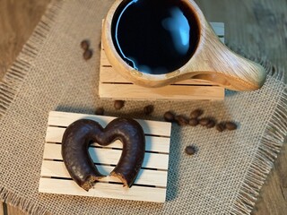 An oak table with visible grains is standing on a wood cup with black coffee. On a jute napkin, coffee beans are scattered and a gingerbread bitten heart lies on the pallet 