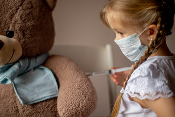 A little girl with pigtails in a mask makes an injection, an inoculation to a big toy bear in a scarf, treats, plays a child's doctor. Health concept, vaccination of children, pediatrician
