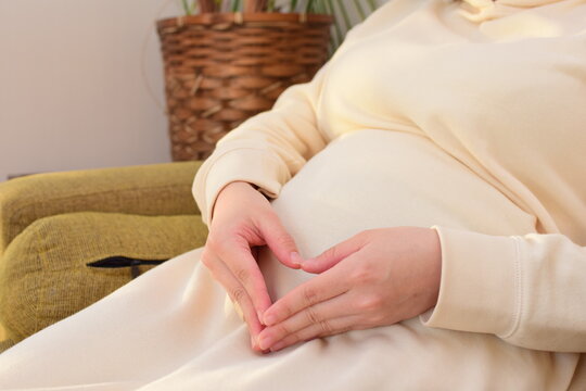 A pregnant woman in her twenties sitting on the sofa is making a heart symbol by hand in front of her big belly.