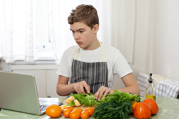 A teenage boy prepares a fruit salad on a recipe from the Internet. The boy cuts lettuce on the kitchen table, in front of him is a laptop. The child is engaged in household chores.