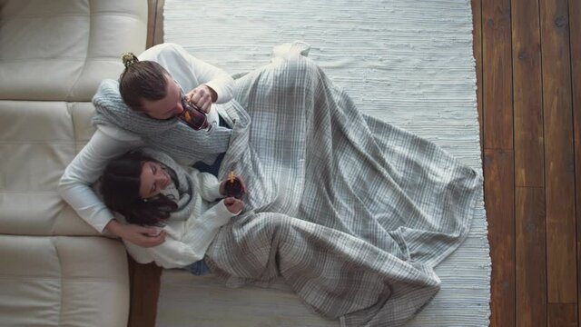 Winter holidays, a couple of young people relax lying on the floor in a country house, man and young woman communicate and drink mulled wine.